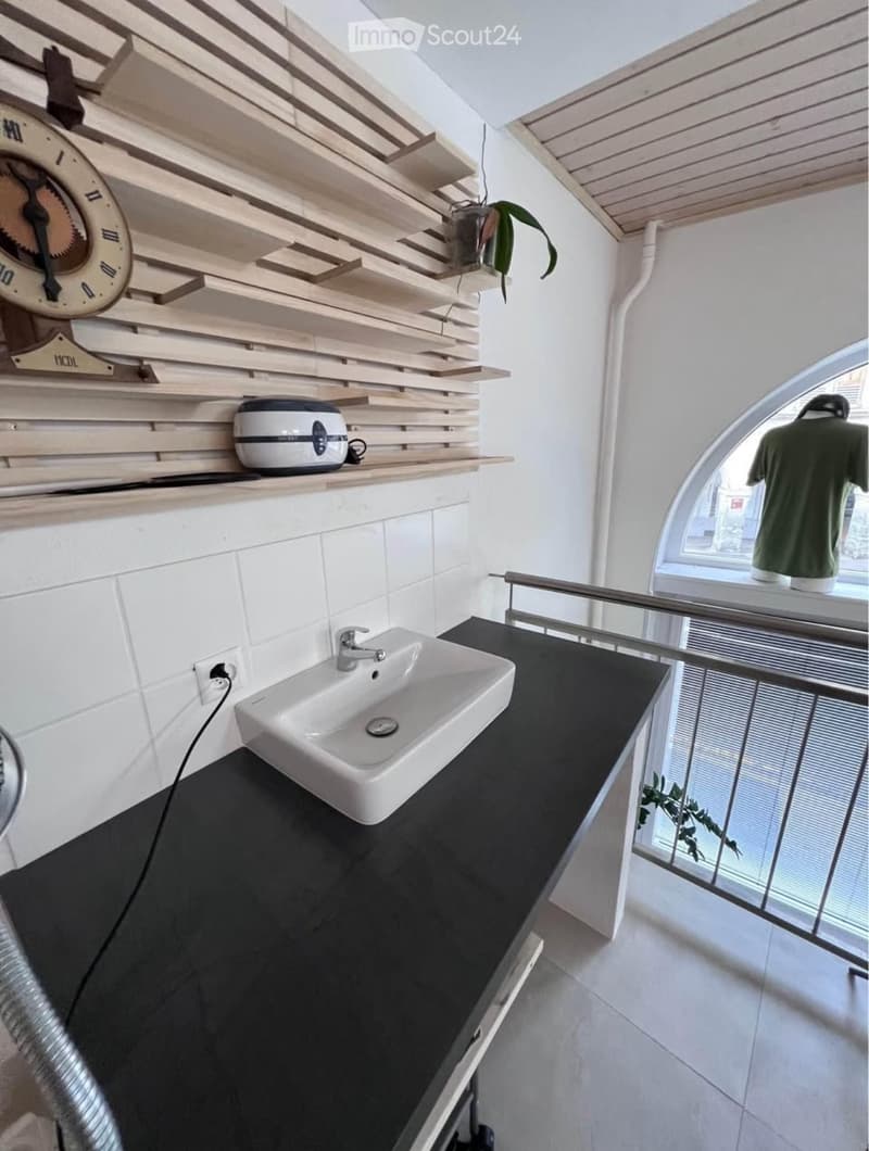 Modern bathroom with white tiled walls, black countertop, white sink, shelves with a clock and toaster, and a plant on the windowsill.