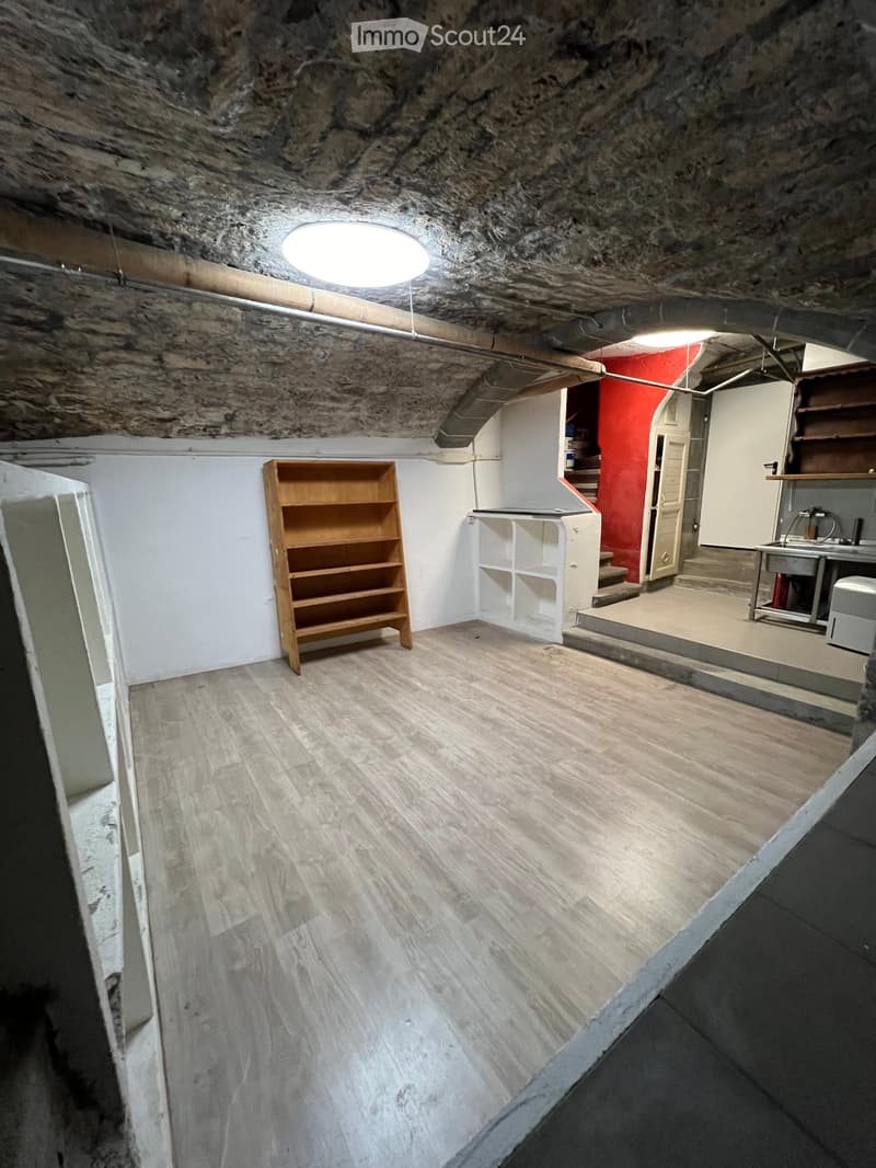 A spacious basement room with wooden flooring, white walls, and exposed stone ceiling. Contains a wooden bookshelf, a white shelf unit, a sink area with a stool, and a staircase leading upwards.