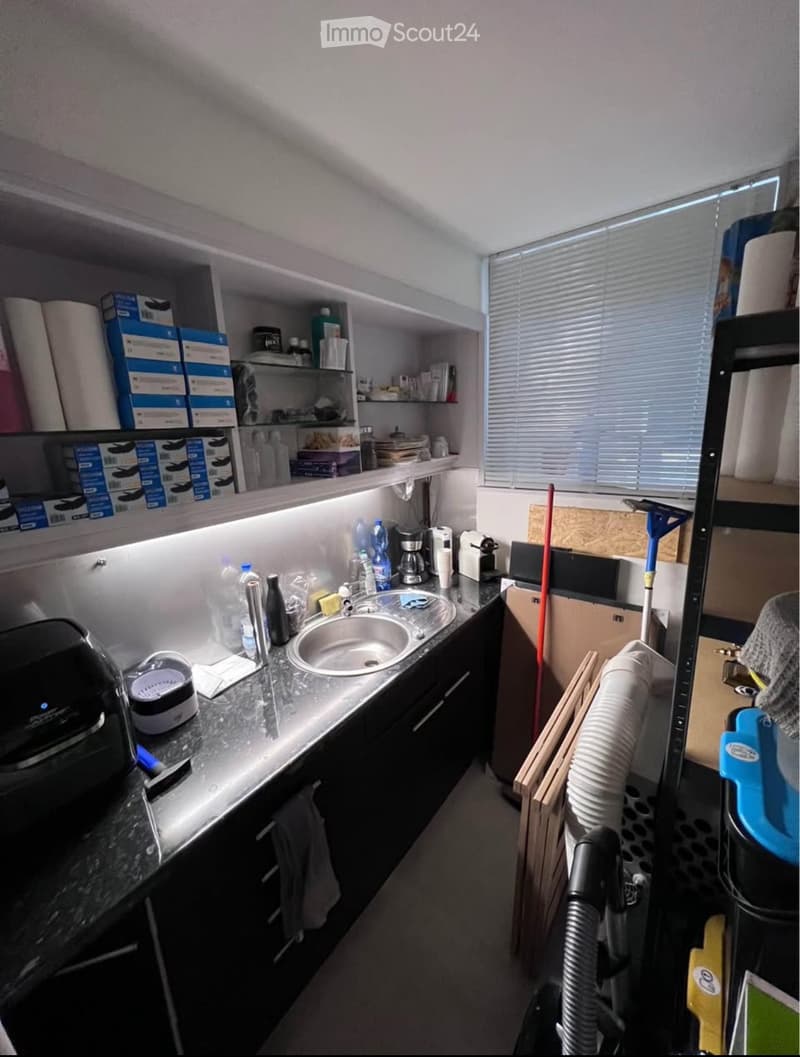 Compact kitchen with a sink, coffee maker, and shelves holding cleaning supplies and boxes. A vacuum cleaner is positioned near the door.