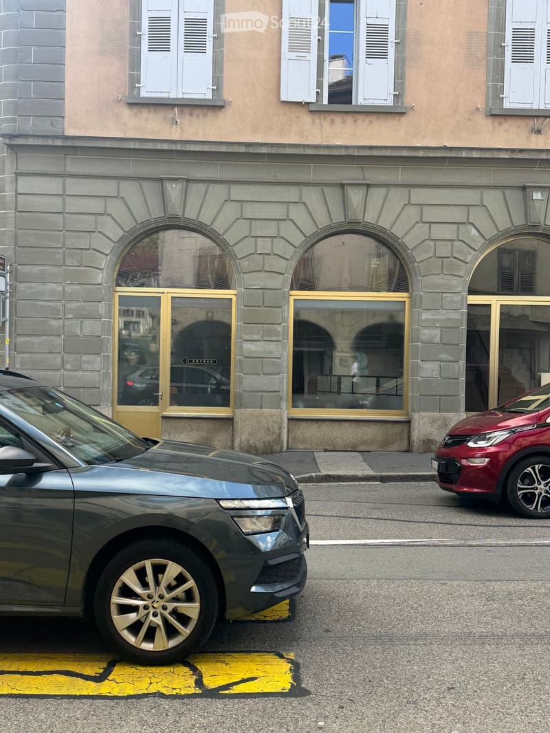 A multi-story building with arched entrances and large windows, featuring a mix of stone and plaster facades. Two cars are parked in front of the building.