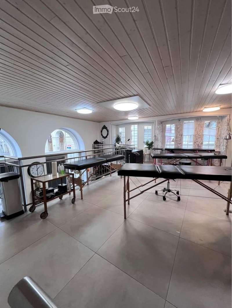 Well-lit room with tiled floor, slanted wooden ceiling, and large windows. Contains treatment tables, a cart with supplies, a mirror, and a clock.