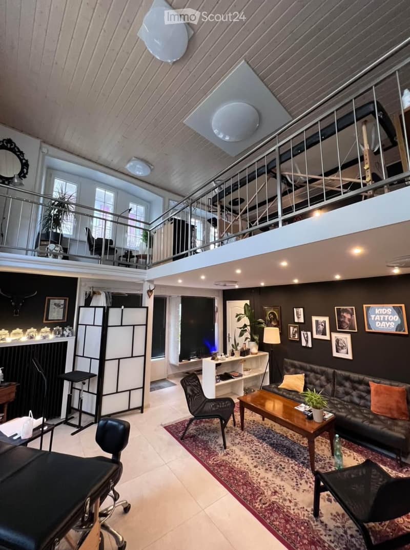 Modern living room with dark walls, exposed wooden ceiling beams, and a lofted upper level. Features a black leather sofa, wooden coffee table, black chairs, a white shelving unit, and decorative items including framed pictures and a sign that reads "Kids