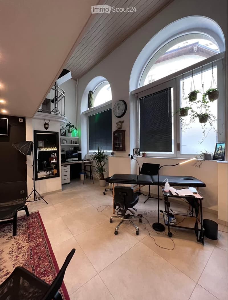 Well-lit room with large arched windows, a massage table, a desk with a laptop, a chair, a stool, a mirror, a lamp, and several plants.
