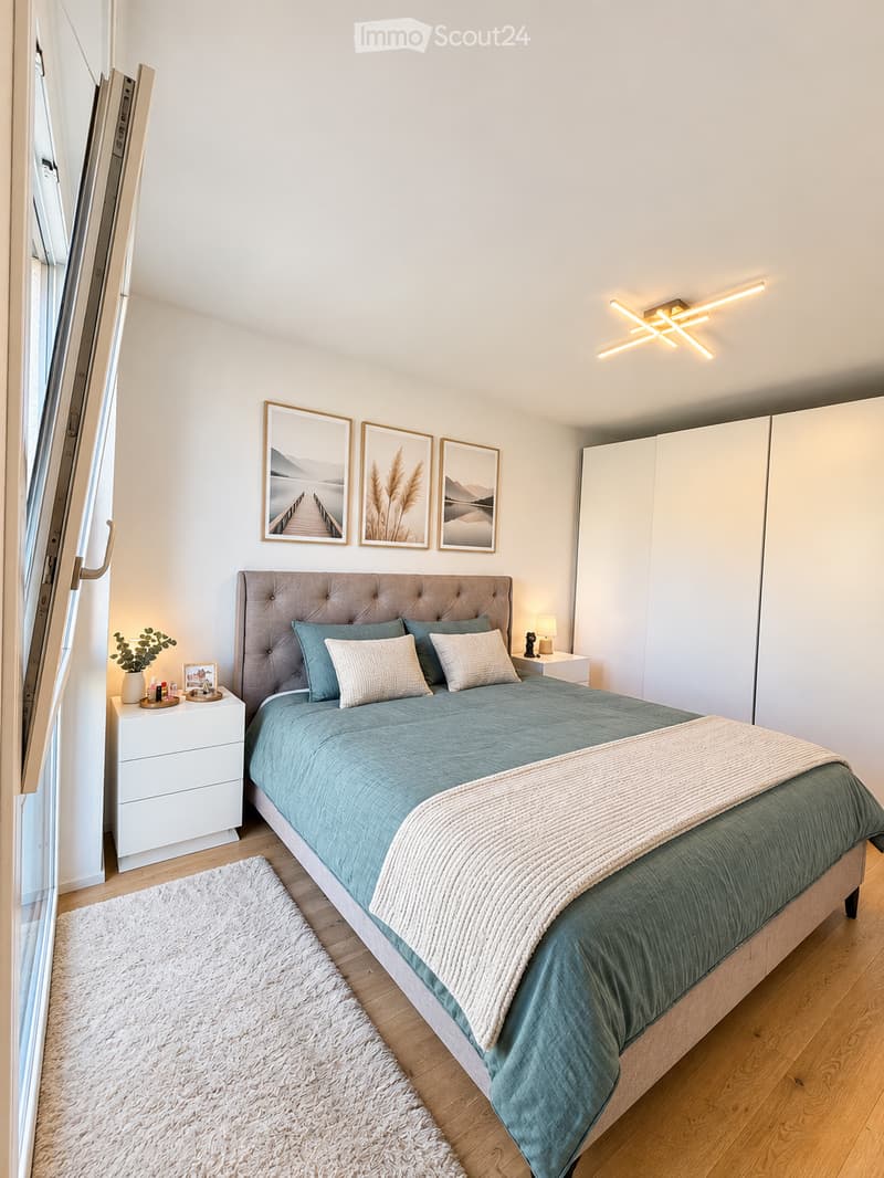 Modern bedroom with a large bed, nightstand, lamp, picture frames, plant, and a white wardrobe.