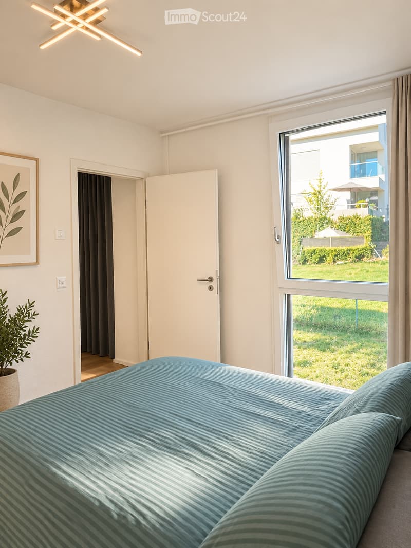 Modern bedroom with a large bed, a potted plant, and a picture frame on the wall. The room has large windows with a view of a green outdoor area.