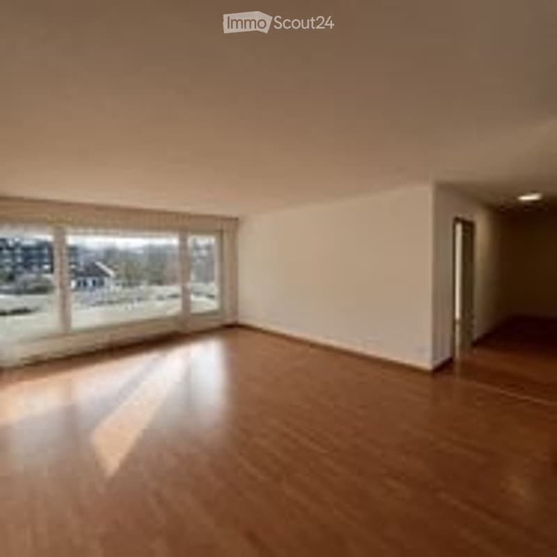 Empty room with wooden flooring, large windows, white walls, and a door leading to another area.