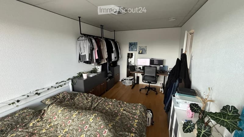 A compact bedroom with a bed, desk, and wardrobe. The room features a wooden floor, a hanging clothes rack, and a small plant on the desk.