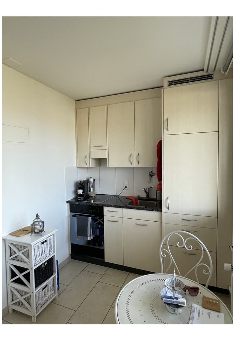 The image shows a modern kitchen with light-colored cabinets, a black oven, a sink with a faucet, and a small dining area with a round table and a chair. There is also a white storage unit with baskets and a small decorative item on top.