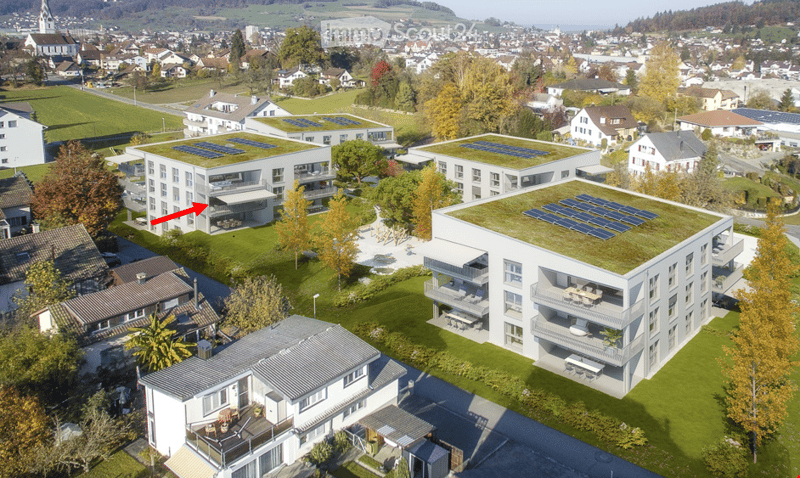 Aerial view of a modern residential complex with multiple buildings, green roofs, and solar panels. The complex is surrounded by a well-maintained lawn and trees, with neighboring houses and a town visible in the background.