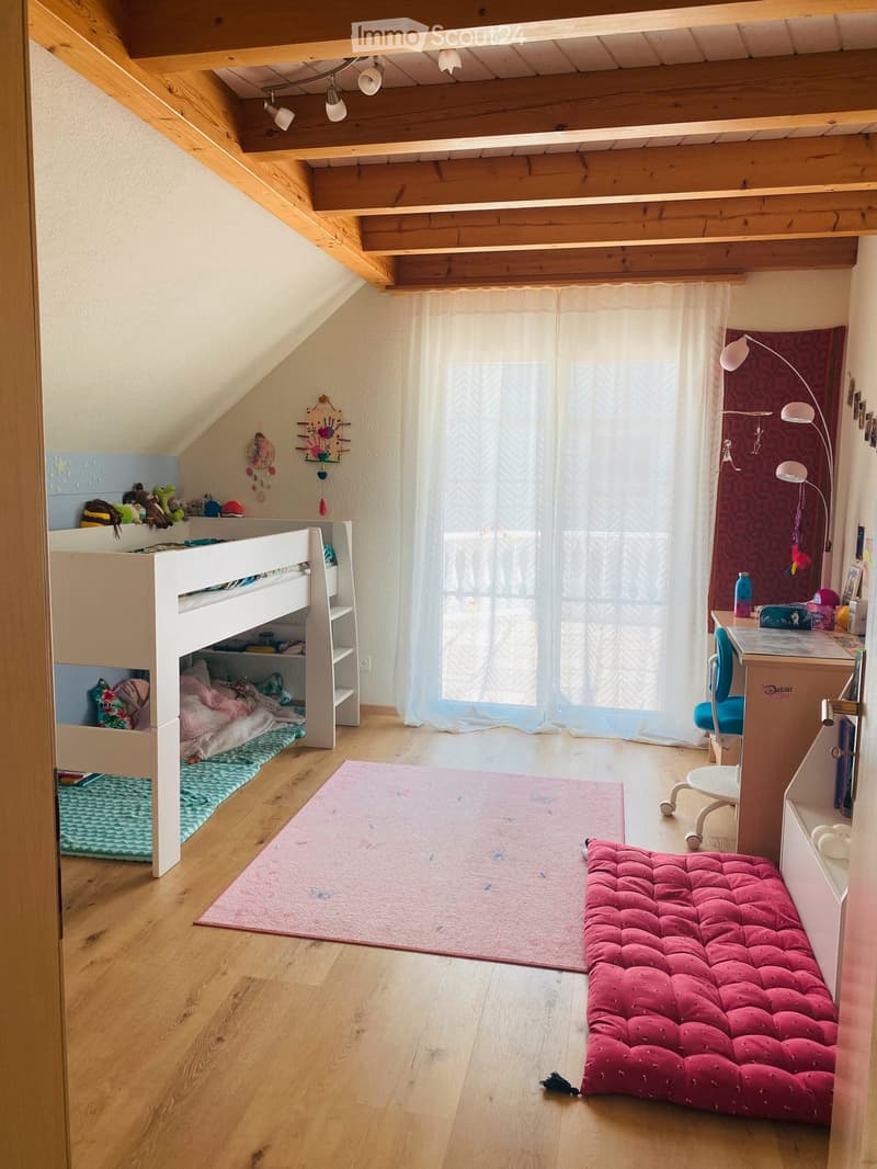 Cozy attic bedroom with wooden slanted ceilings, bunk bed, desk, and a red tufted bench. Features a large window with sheer curtains and a colorful rug.