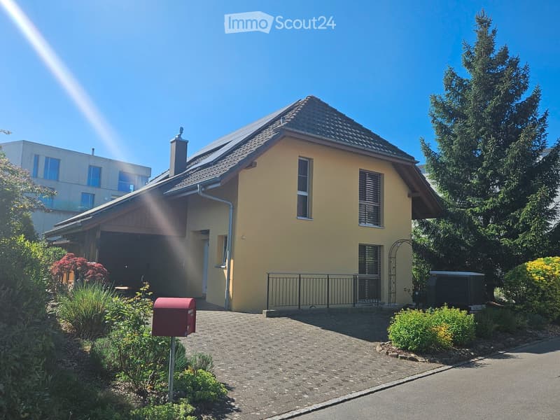 Single-story house with a yellow exterior, dark roof, and a small garage. The house has multiple windows, a chimney, and is surrounded by greenery and a well-maintained garden.