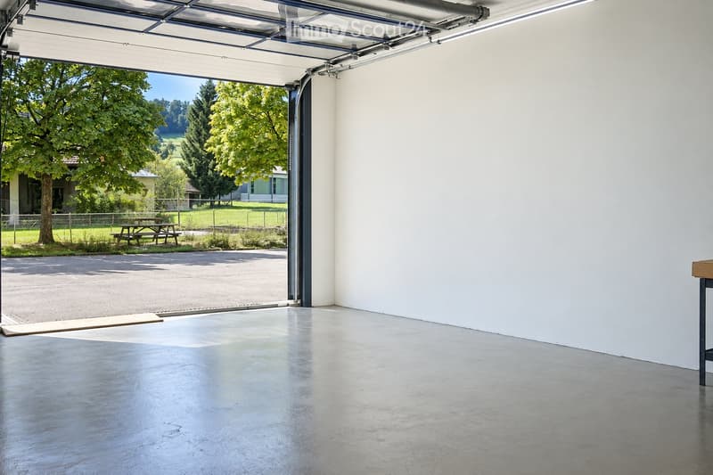Empty garage with a large, open garage door leading to a grassy area with trees and a picnic table outside.