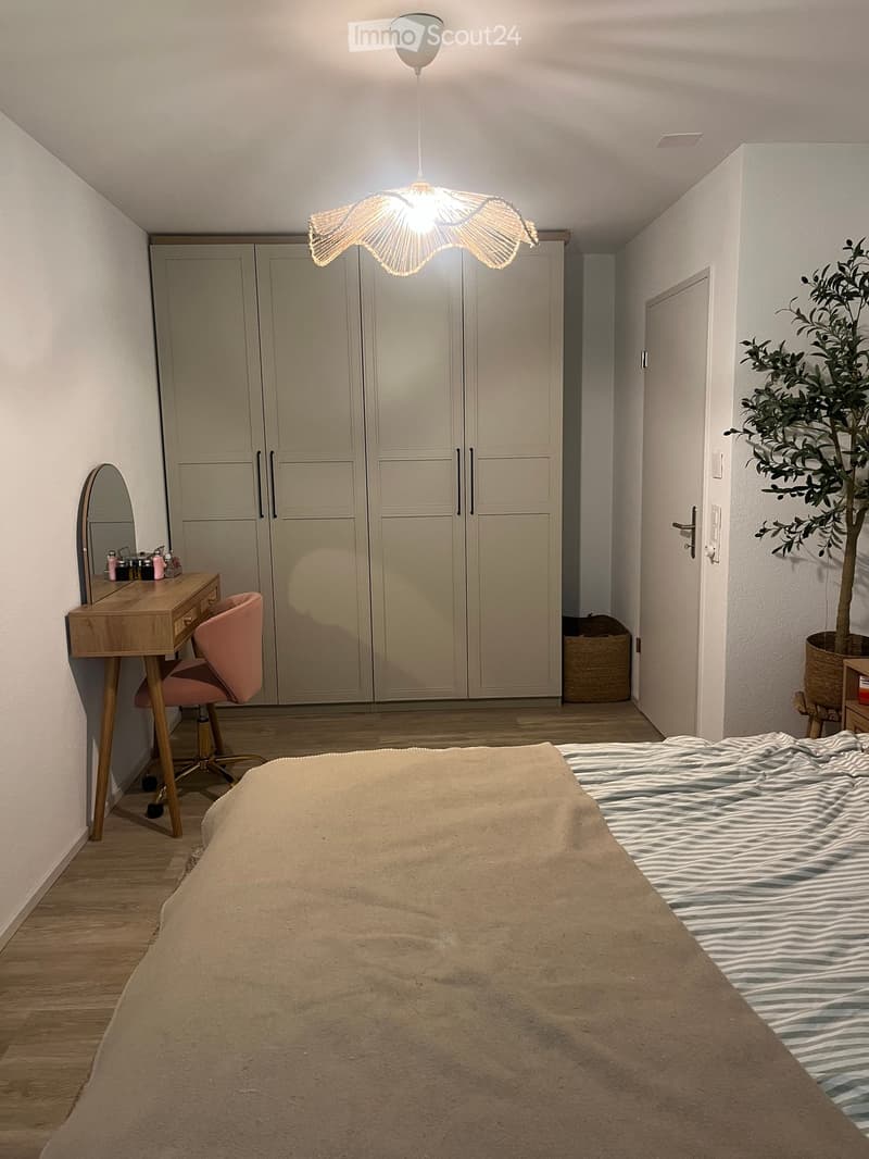 Modern bedroom with a large bed, a pink chair, a wooden desk with a mirror, a wardrobe, and a hanging lamp.