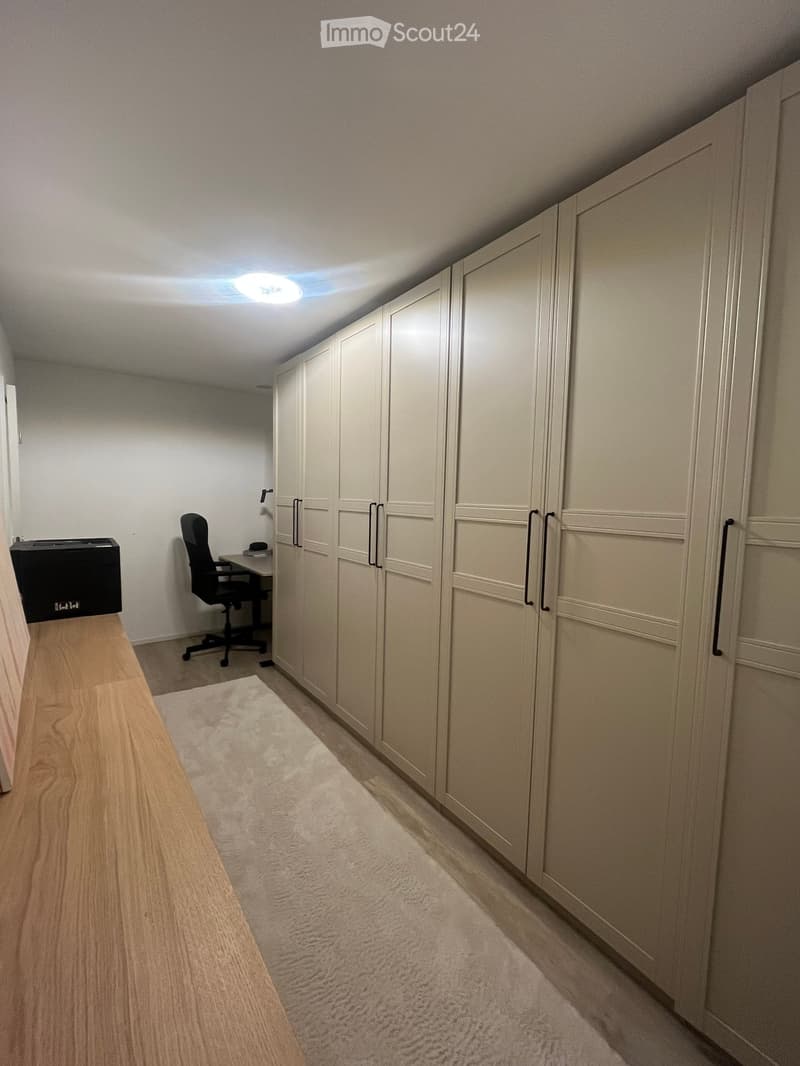 A small, neatly organized room with built-in wardrobes, a desk, a chair, and a printer. The room features a carpeted floor and recessed ceiling lighting.
