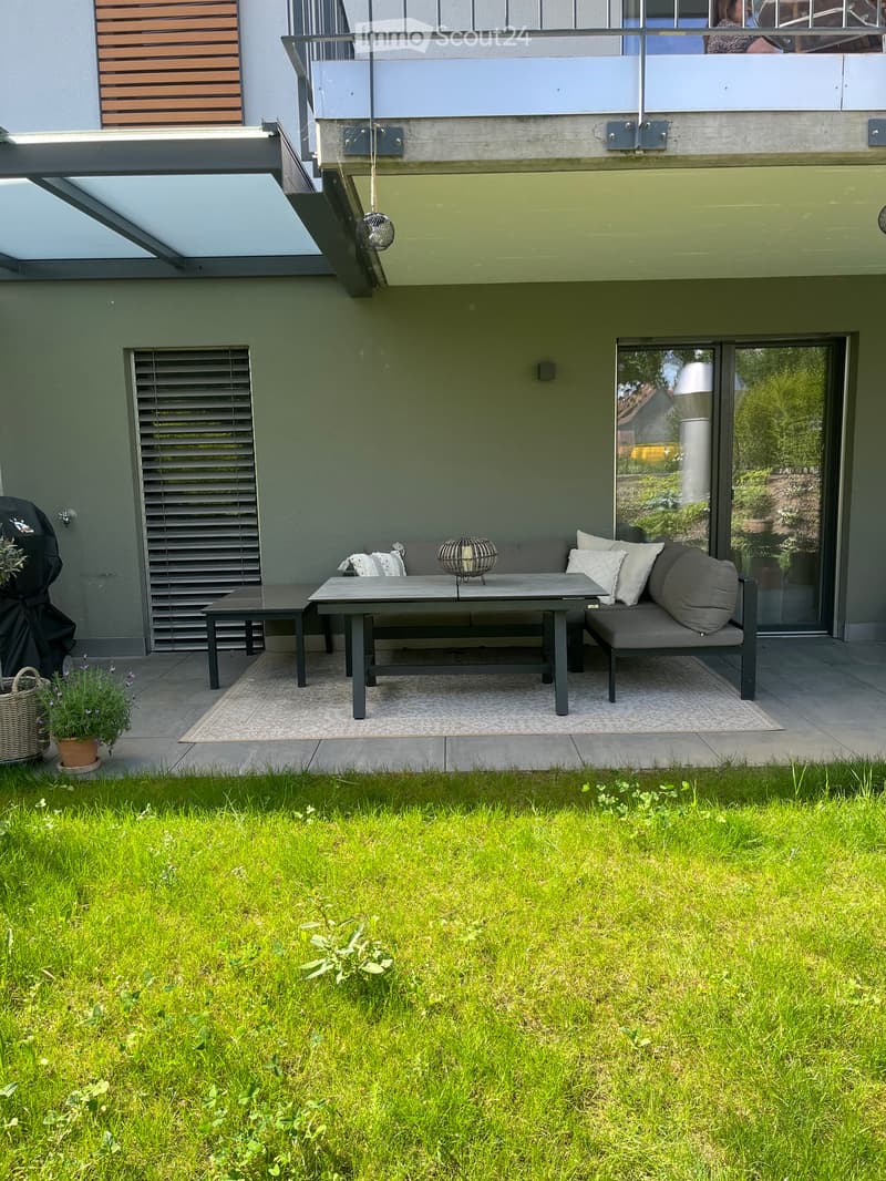 Modern outdoor patio with a grey sofa, table, bench, and small garden area with grass and potted plants.
