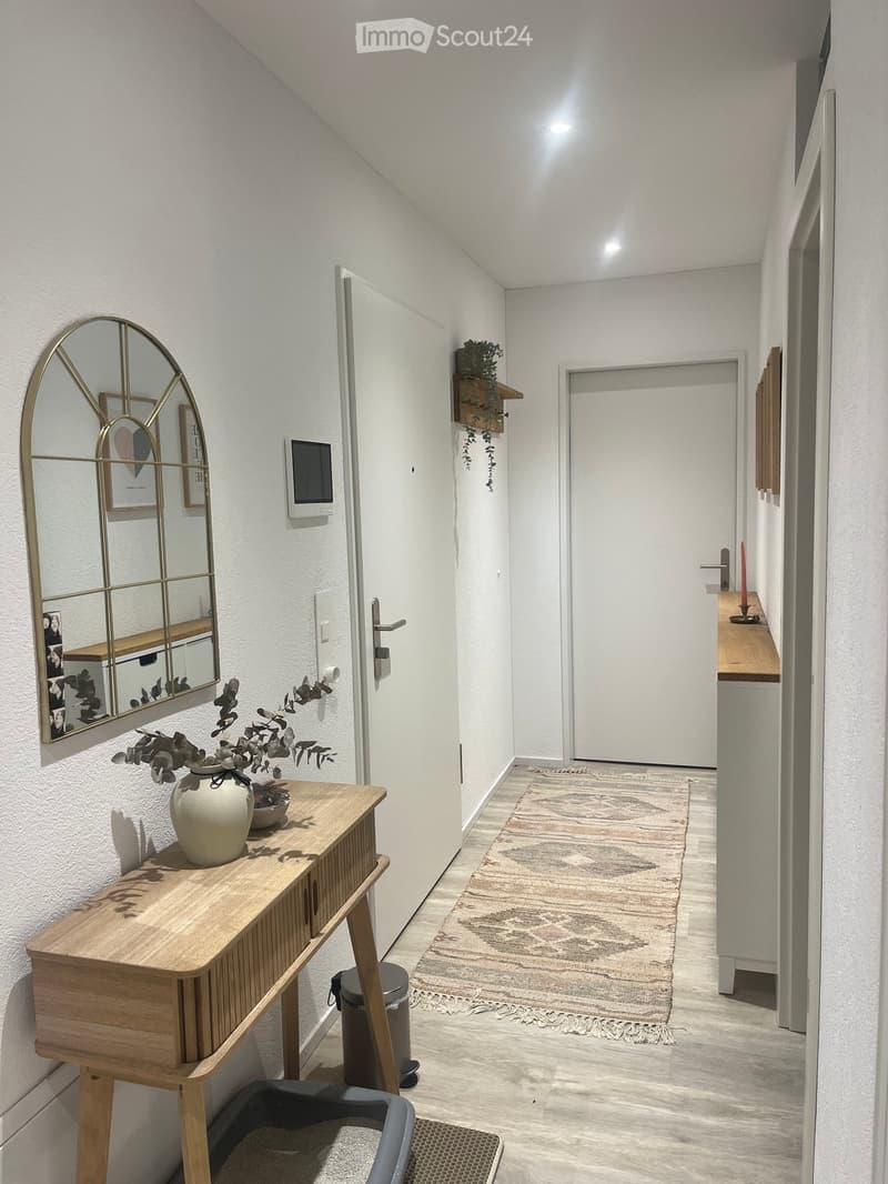 Modern hallway with white walls, wooden floor, and a small rug. Features a wooden console table with a vase, a mirror, and a small shelf with plants.