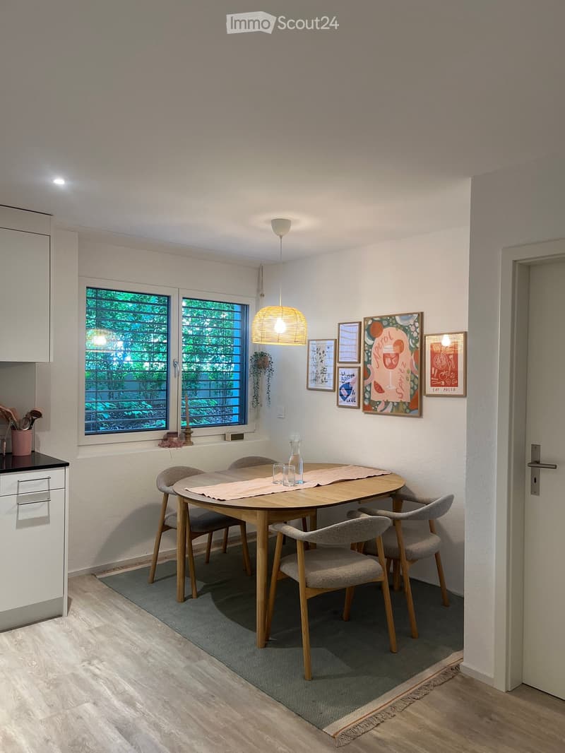 A modern dining area with a round wooden table, four grey chairs, a hanging pendant light, and a white cabinet. The space features large windows with a view of greenery, a grey rug, and several framed pictures on the wall.