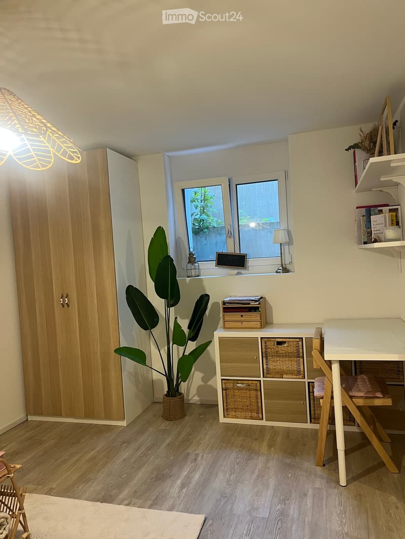 A well-lit room with wooden flooring, a large window with a view of greenery, a tall potted plant, a white desk with a chair, a wooden wardrobe, and a storage unit with woven baskets.