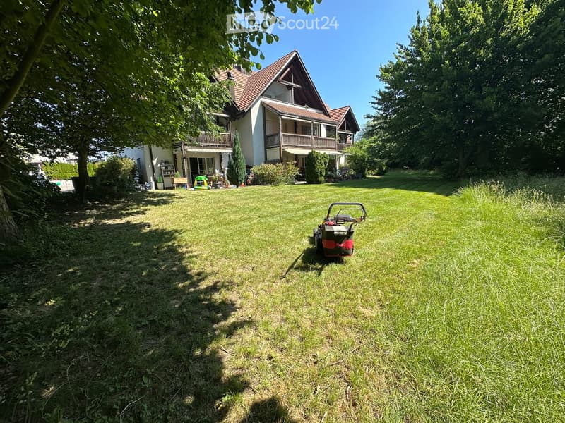 A well-maintained lawn in front of a two-story house with balconies, surrounded by trees and shrubs. A lawnmower is visible on the grass.