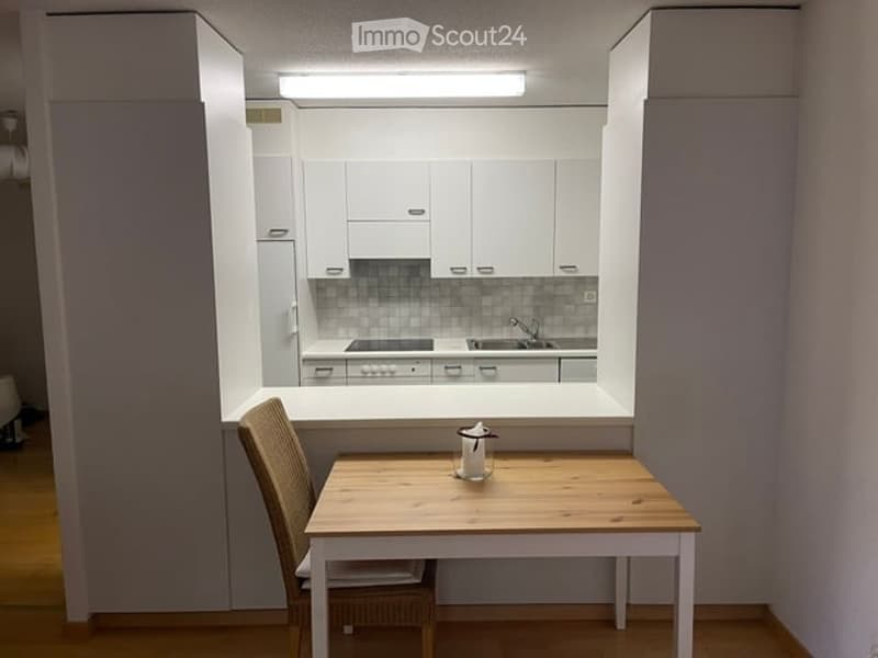 A modern, compact kitchen with white cabinets, a small wooden dining table, and a wicker chair. The kitchen features a sink, stove, and a built-in window for natural light.