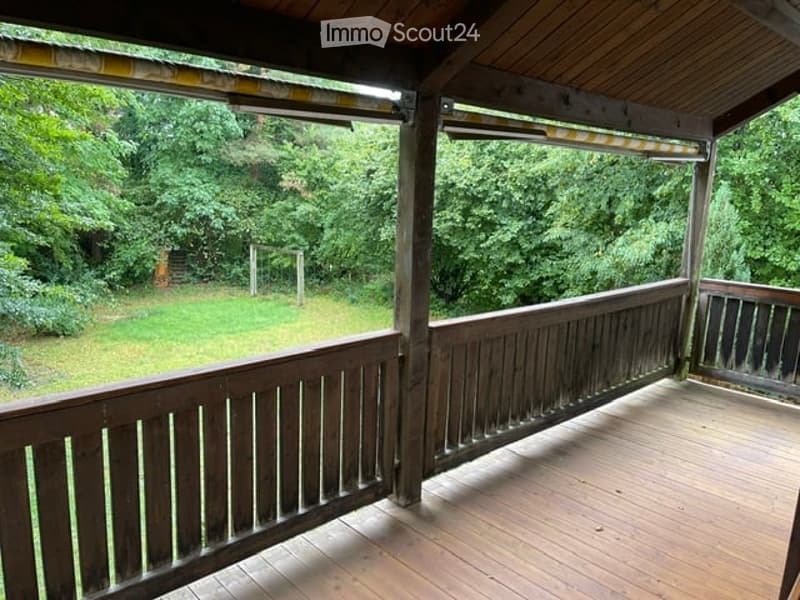Wooden balcony with a wooden railing and a wooden floor, overlooking a green, grassy yard with trees.