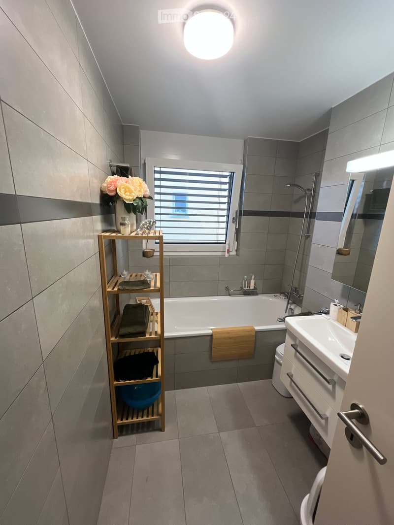 Modern bathroom with grey tiles, a bathtub, sink, toilet, wooden shelf with towels and decorative items, and a window with blinds.