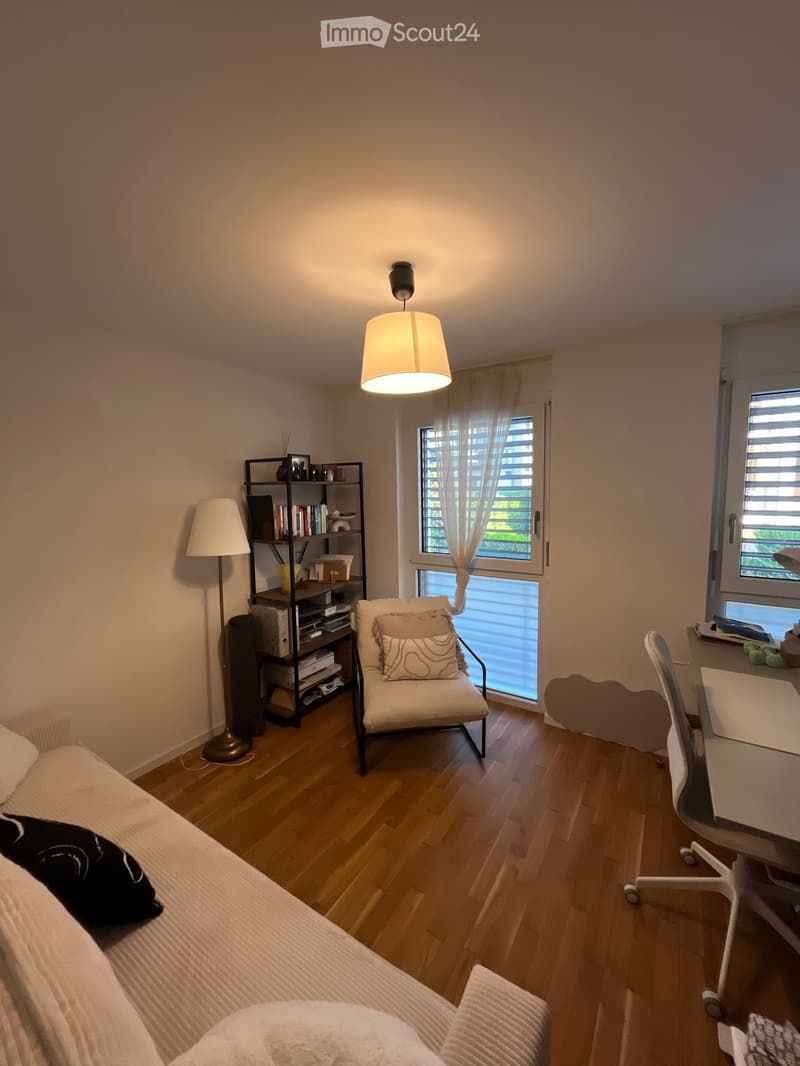Cozy bedroom with wooden flooring, a bed with pillows, a chair, a shelf with books and decor, a desk with a chair, and a hanging lamp.