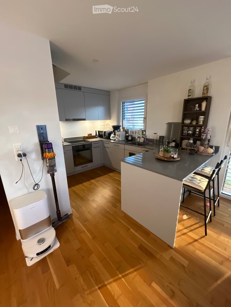 Modern kitchen with wooden flooring, grey cabinets, and a black countertop. Appliances include an oven, microwave, coffee maker, and fridge. There are stools near the island and various kitchen items on the shelves and countertop.