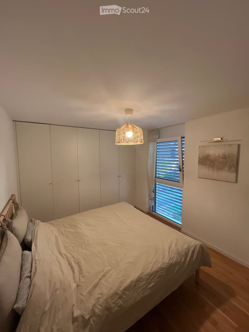 A well-lit bedroom with a large bed, a built-in wardrobe, a window with blinds, and a hanging lamp.