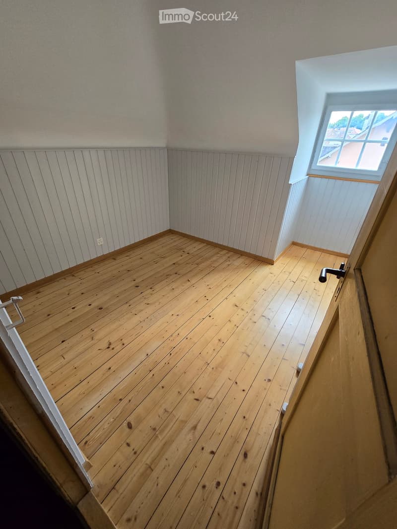 Empty room with wooden floor and white paneled walls, featuring a small window with a view of the outside.