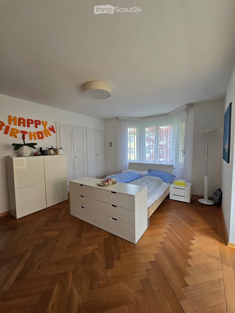 Modern bedroom with wooden flooring, white furniture including a bed, nightstand, wardrobe, and drawer unit. Features a floor lamp, potted plant, and decorative items on a small table.
