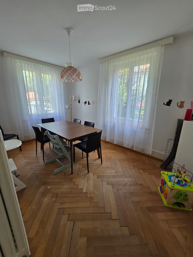 Dining room with wooden floor, large windows with white curtains, a modern dining table with chairs, a colorful toy box, and decorative wall hangings.