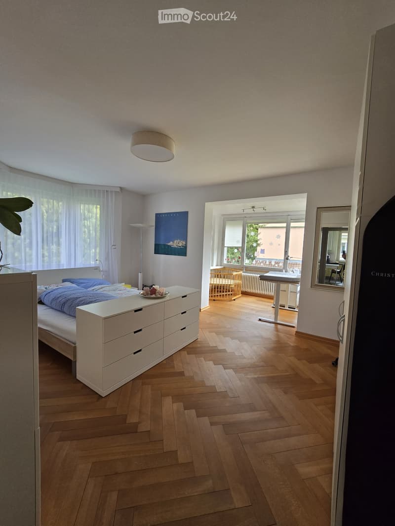 Bright bedroom with wooden flooring, a single bed with blue bedding, a nightstand, a mirror, a small table, a chair, a wooden cradle, and a painting on the wall. The room has large windows with sheer curtains and a view of greenery outside.