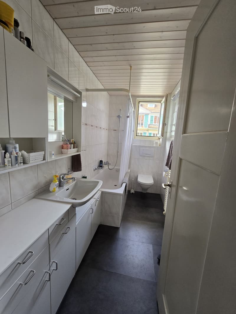 Modern bathroom with white cabinets, a sink, bathtub, toilet, and a window providing natural light. The space features a dark grey tiled floor and white tiled walls.