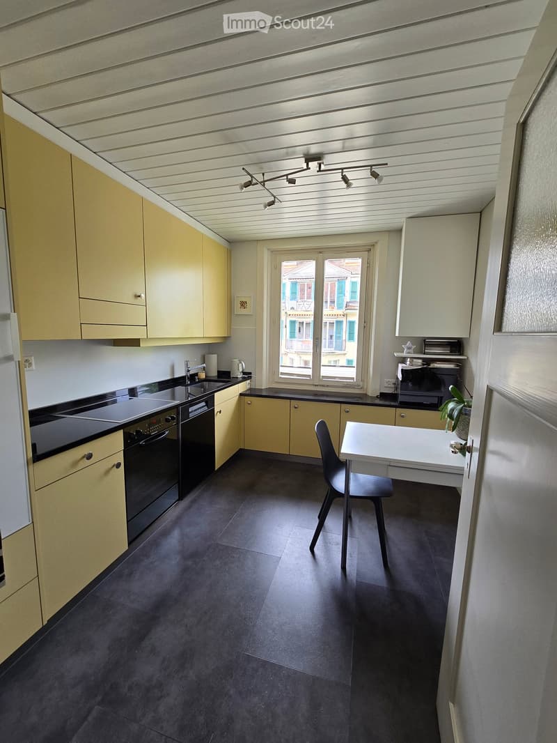 Modern kitchen with light yellow cabinets, black countertops, and a dark tiled floor. Features include a sink, oven, small table with a chair, and a potted plant on the counter.