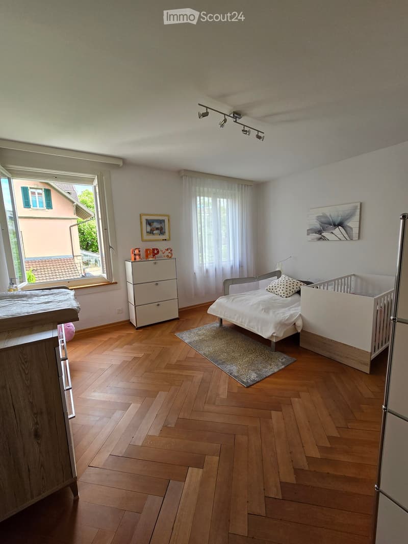 Brightly lit bedroom with wooden flooring, a single bed, a crib, a small cabinet, and a large window with white curtains.