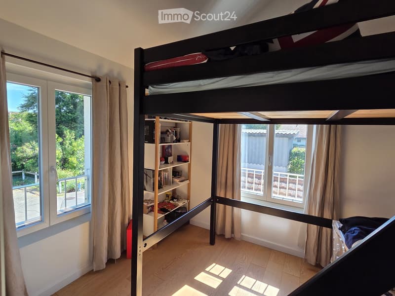A cozy bedroom with a lofted bed, built-in shelving unit, and large windows with curtains, allowing natural light to fill the space.