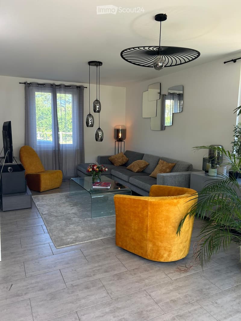 Modern living room with grey sofa, glass coffee table, mustard yellow chairs, and decorative mirrors on the wall. Features pendant lights and a large window with sheer curtains.