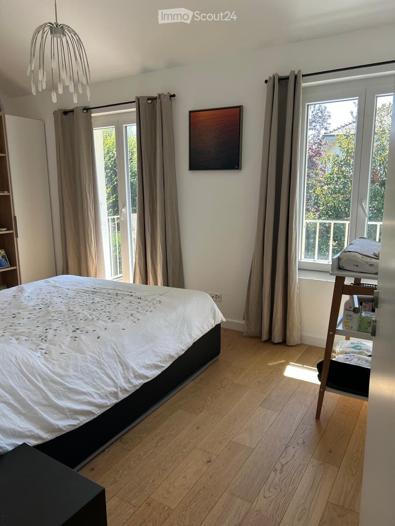 Bright bedroom with wooden flooring, large windows, and a bed with white bedding. Features include a modern chandelier, a painting on the wall, and a nightstand with items.