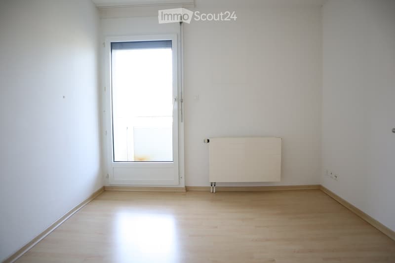 Empty room with white walls, wooden floor, large window with blinds, radiator, and electrical outlet.