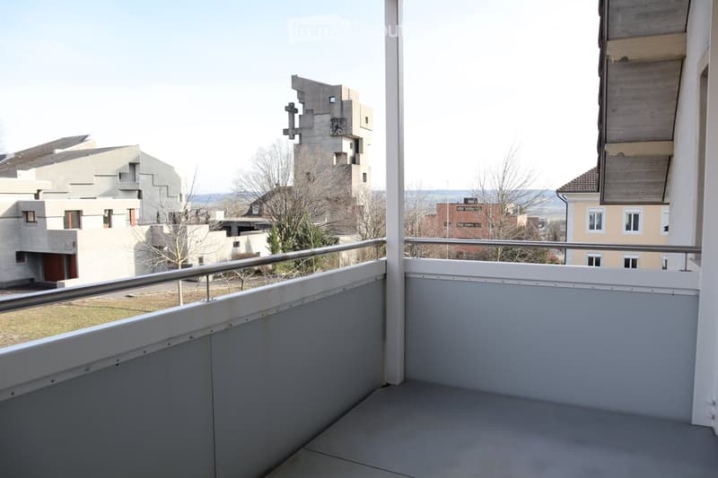 Balcony with white railing and grey floor, overlooking a modern residential area with contemporary buildings and a distant landscape.