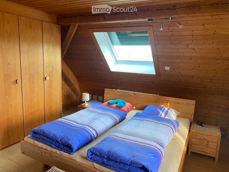 Cozy attic bedroom with wooden walls and slanted ceilings, featuring a double bed with blue and red striped bedding, a nightstand with a lamp, and a large skylight window.