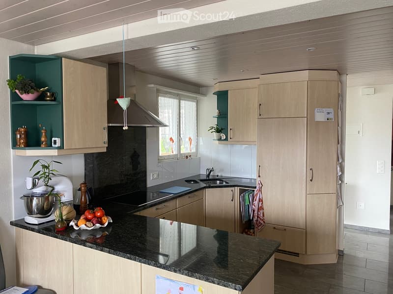 Modern kitchen with light wooden cabinets, granite countertops, and stainless steel appliances. Features include a central island, pendant lighting, and built-in storage shelves.