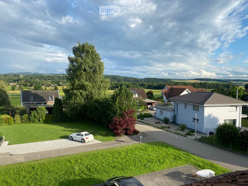 A modern two-story house with a white exterior, surrounded by a well-maintained lawn and trees. The driveway features a white sports car and a black car. The house has a balcony on the upper level and is situated in a rural area with other houses and gree