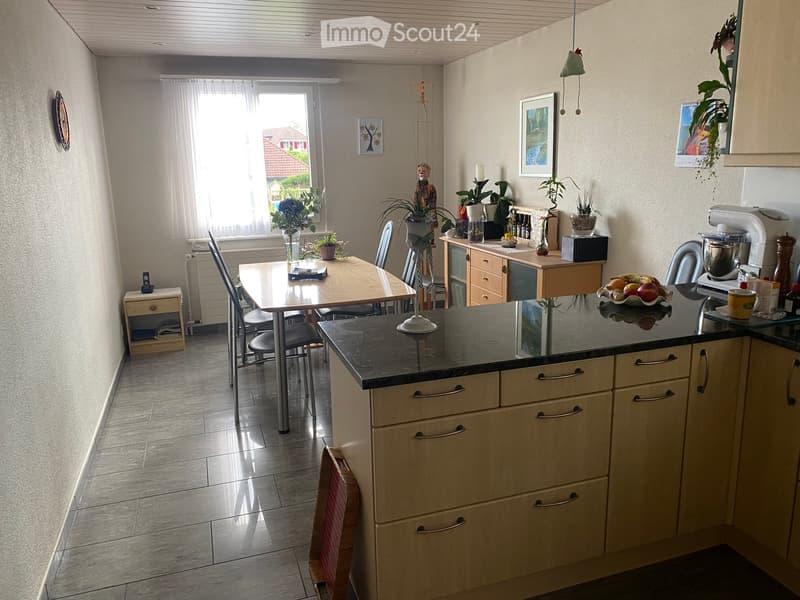 Bright kitchen with a dining area, featuring a dark countertop, light wooden cabinets, a mixer, and a bowl of fruit on the counter.