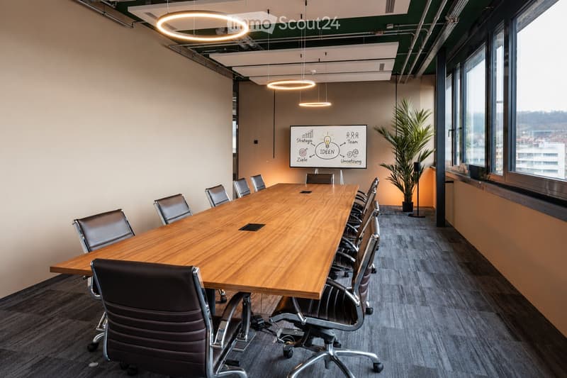 Modern conference room with a long wooden table surrounded by black leather chairs, a large screen on the wall, and a potted plant near the window.