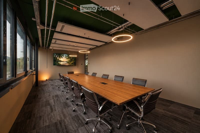 Modern conference room with a long wooden table, multiple chairs, and a large screen on the wall. The room features large windows, pendant lights, and a neutral color palette.
