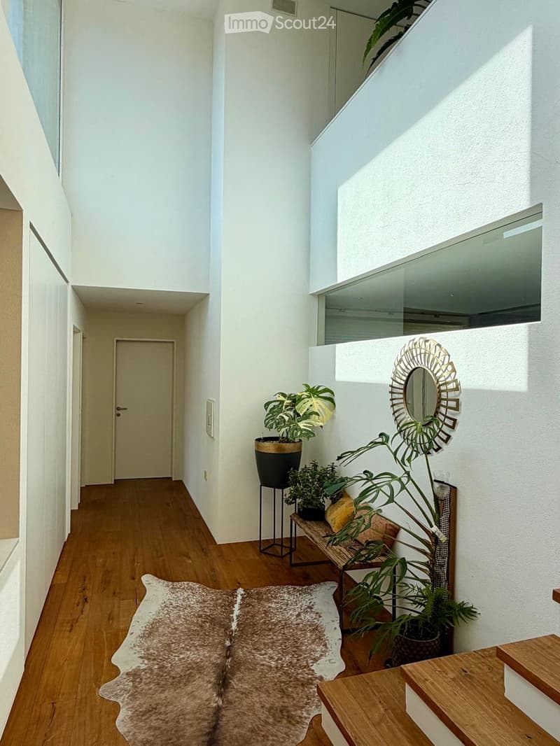 Modern hallway with wooden flooring, white walls, and natural light from skylights. Features include a small bench with cushions, a cowhide rug, potted plants, and a decorative mirror on the wall.