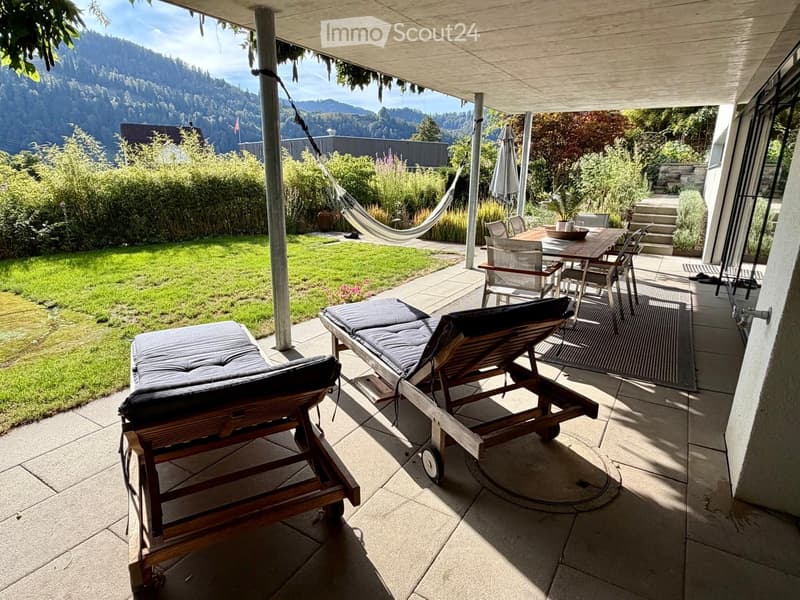 A covered outdoor patio with lounge chairs, a dining table, and a view of a green lawn and mountains.