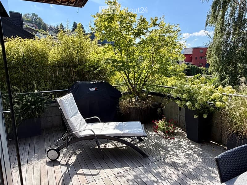 Balcony with wooden decking, lounge chair, covered grill, and lush greenery in the background.